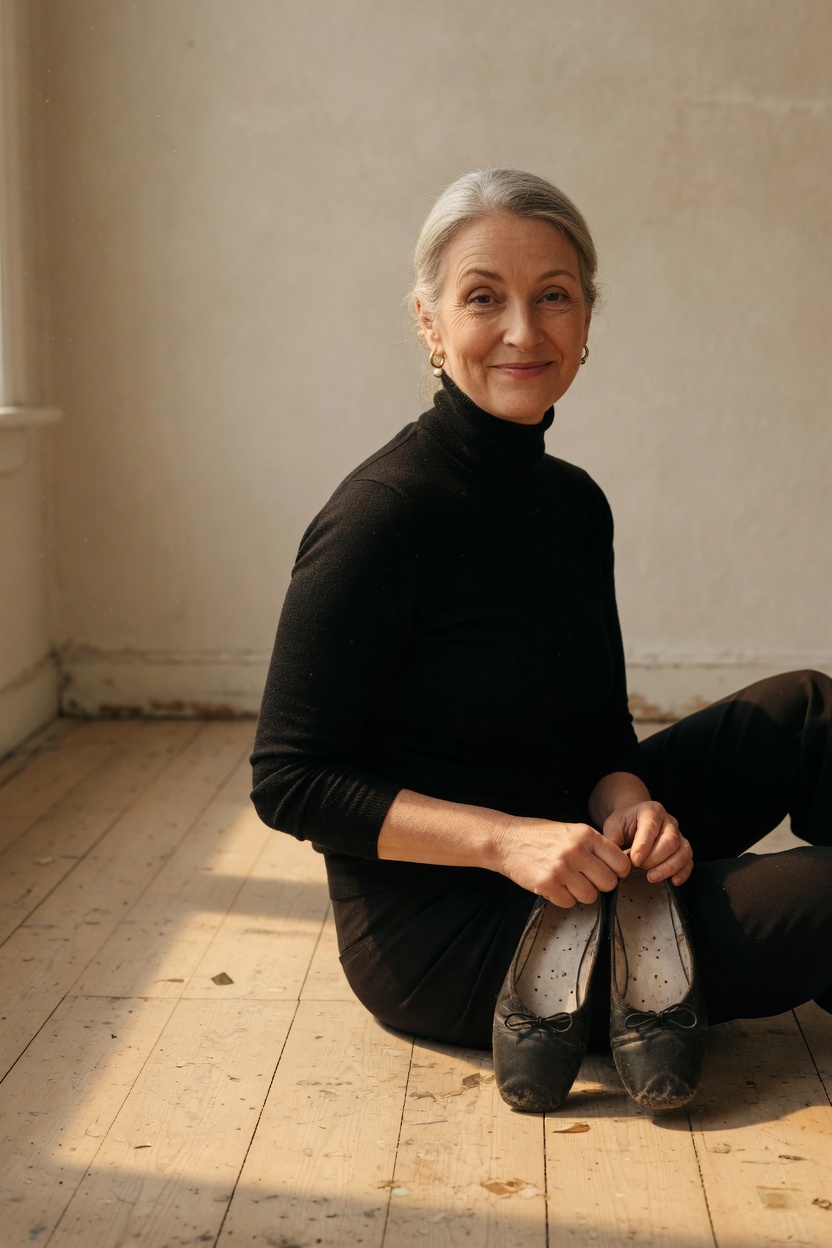 Sandra in a black turtleneck, silver hair in a low bun, worn ballet slippers in her hands, seated on a wooden floor.